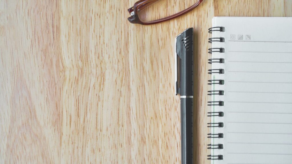 机にボールペン・ノート・茶色のメガネが置いてある画像